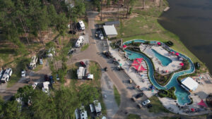 Aerial drone view of Fireside RV Resort showing the winding lazy river, swimming pools, RVs parked among tall pine trees, and the resort's lake