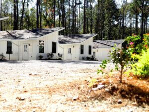 Row of modern white cabins with dark trim at Fireside RV Resort lined along a gravel drive backed by tall Louisiana pines on a sunny day