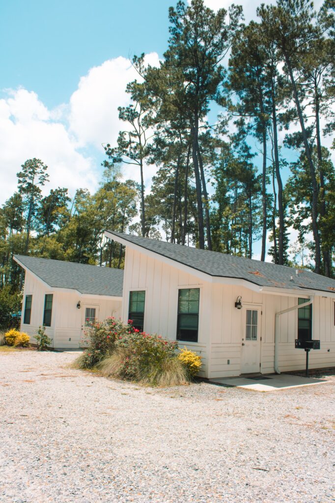 Two modern white cabins with dark shingled roofs and vertical siding at Fireside RV Resort, surrounded by tall Louisiana pine trees under a bright blue sky