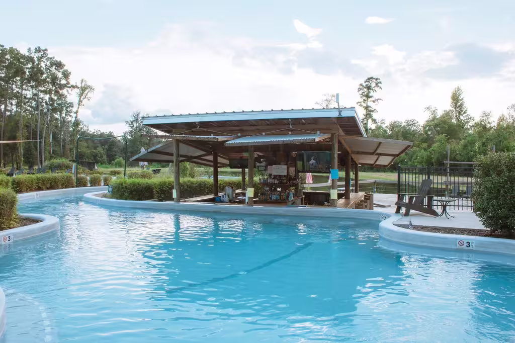A wooden, open-air swim-up bar with a metal roof sits in the center of a winding lazy river at an outdoor resort. The bright blue water curves around at Fireside RV Resort in Ponchatoula, Louisiana.