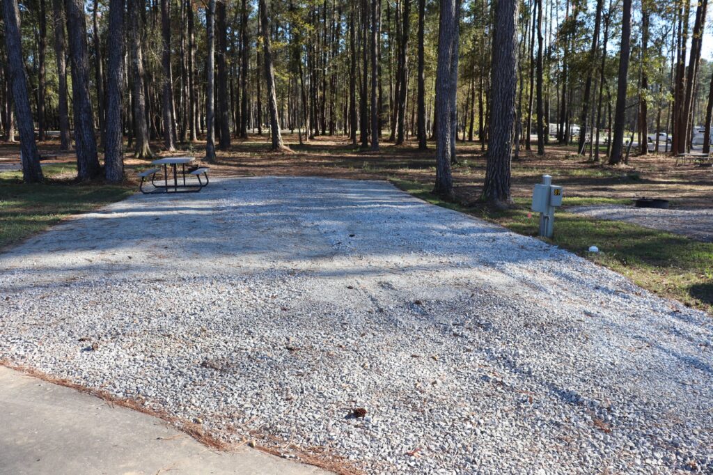 A shaded gravel RV site at Fireside RV Resort surrounded by tall pine trees, with picnic table and electrical hookup pedestal