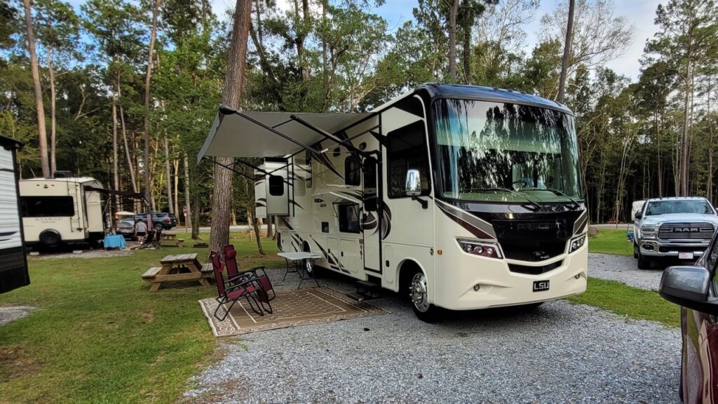 A Class A motorhome with its awning extended over chairs and an outdoor rug on a shaded gravel RV site at Fireside RV Resort in Ponchatoula, Louisiana