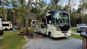 A Class A motorhome with its awning extended over chairs and an outdoor rug on a shaded gravel RV site at Fireside RV Resort in Ponchatoula, Louisiana