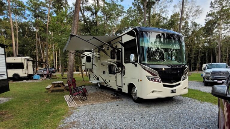 A Class A motorhome with its awning extended over chairs and an outdoor rug on a shaded gravel RV site at Fireside RV Resort in Ponchatoula, Louisiana