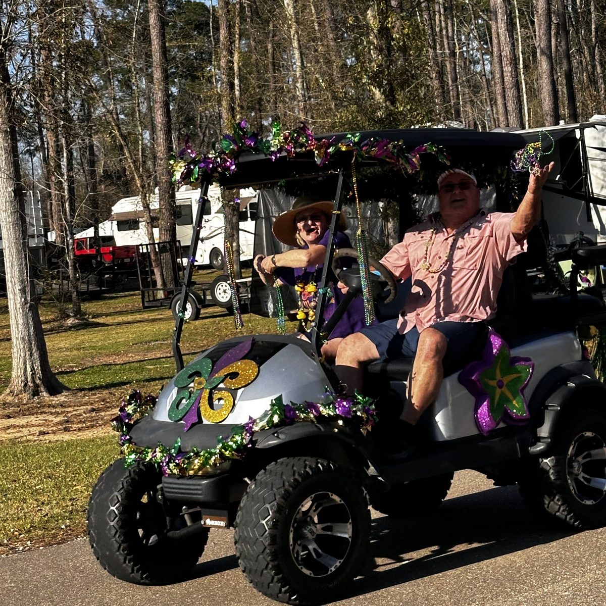 Man in a pink shirt waving beside a woman in a straw hat on a silver golf cart decorated for Mardi Gras at Fireside RV Resort in Ponchatoula