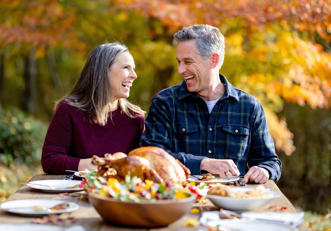A man and a woman laughing over a roast turkey at an outdoor Thanksgiving table on a mild autumn afternoon