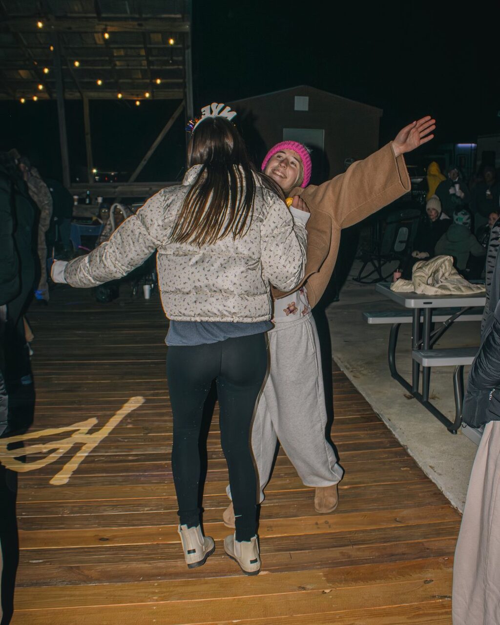 Two guests in winter jackets and a pink beanie dancing on the wooden deck at night at Fireside RV Resort near New Orleans