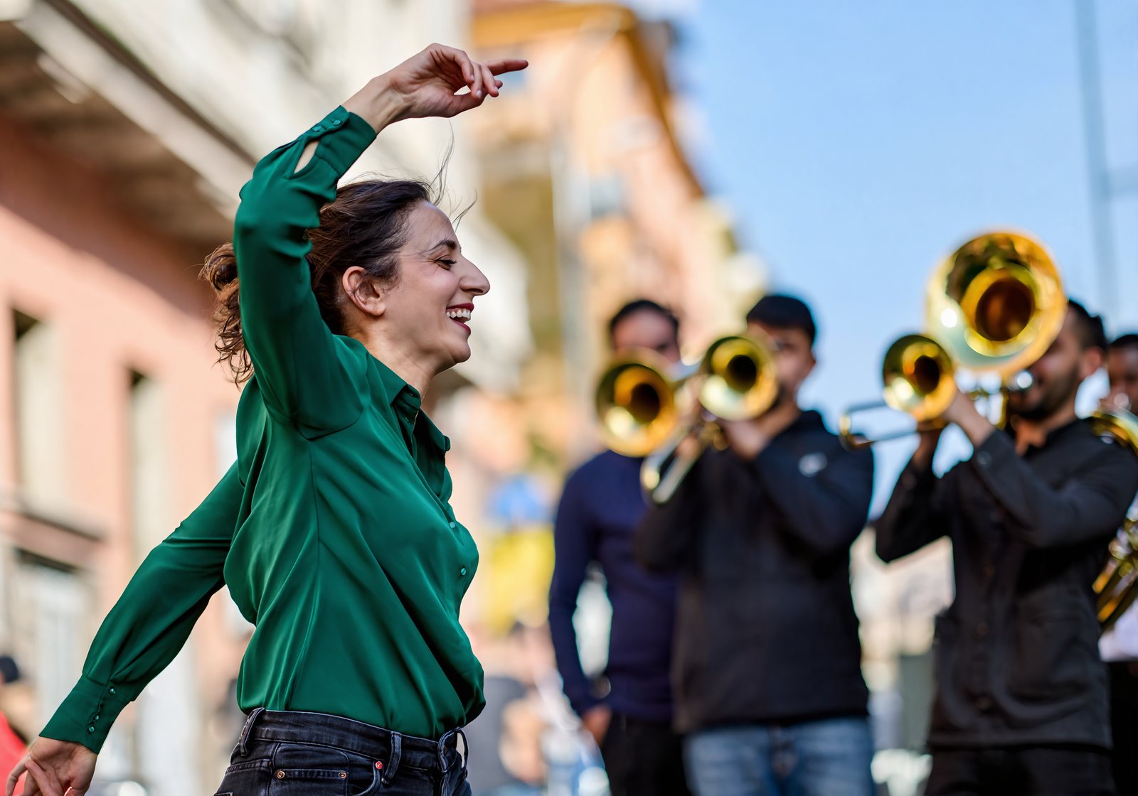 A woman in a green blouse dances in the street as a brass band plays behind her on a sunny afternoon