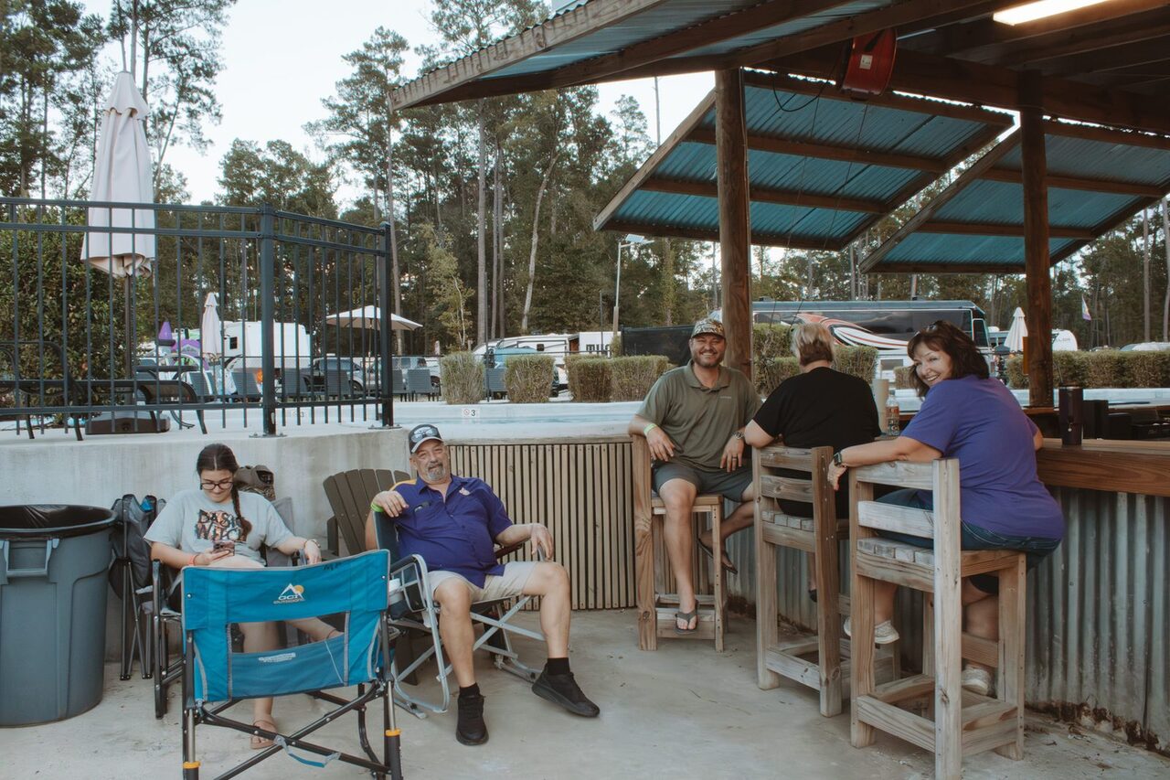 Guests chatting on wooden stools and camp chairs in a shaded gathering area at Fireside RV Resort near Ponchatoula