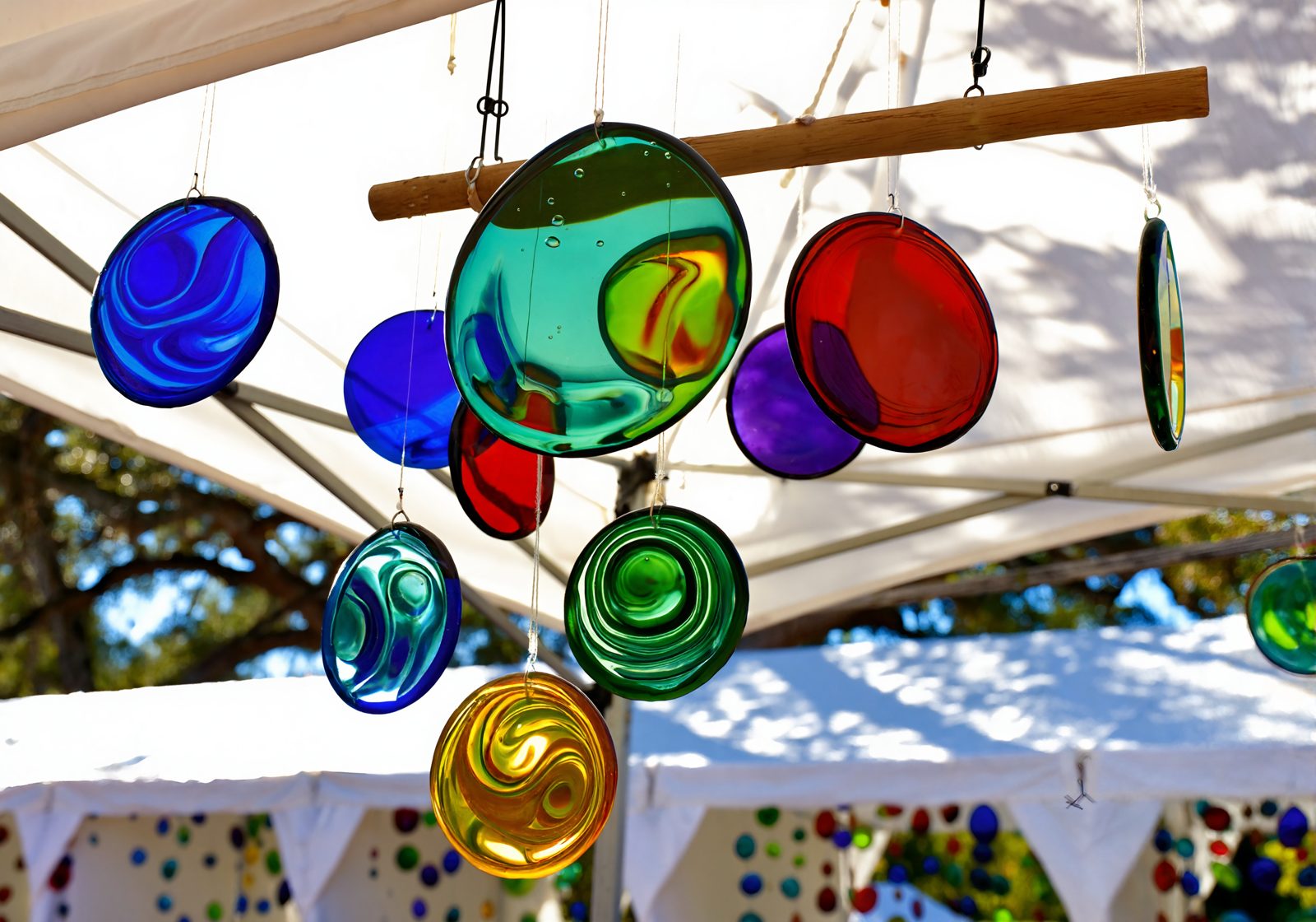 Colorful handmade fused-glass suncatchers hanging in sunlight inside a white booth at an outdoor fall art festival