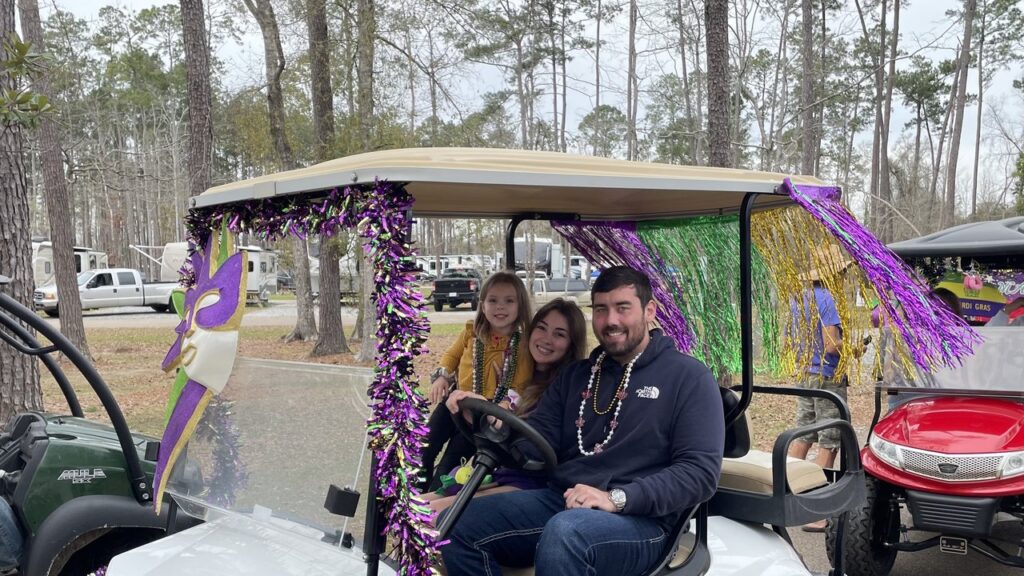 Family riding a golf cart decorated in purple, green and gold Mardi Gras tinsel past RVs under tall pines at Fireside, a seasonal RV park in Louisiana