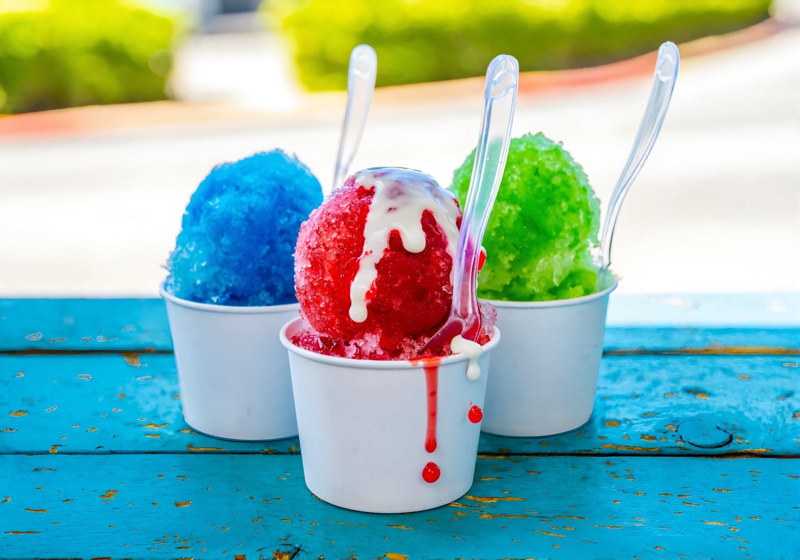 Three sno-balls of shaved ice with red, blue and green syrup in white cups, one topped with condensed milk, on a blue wooden counter
