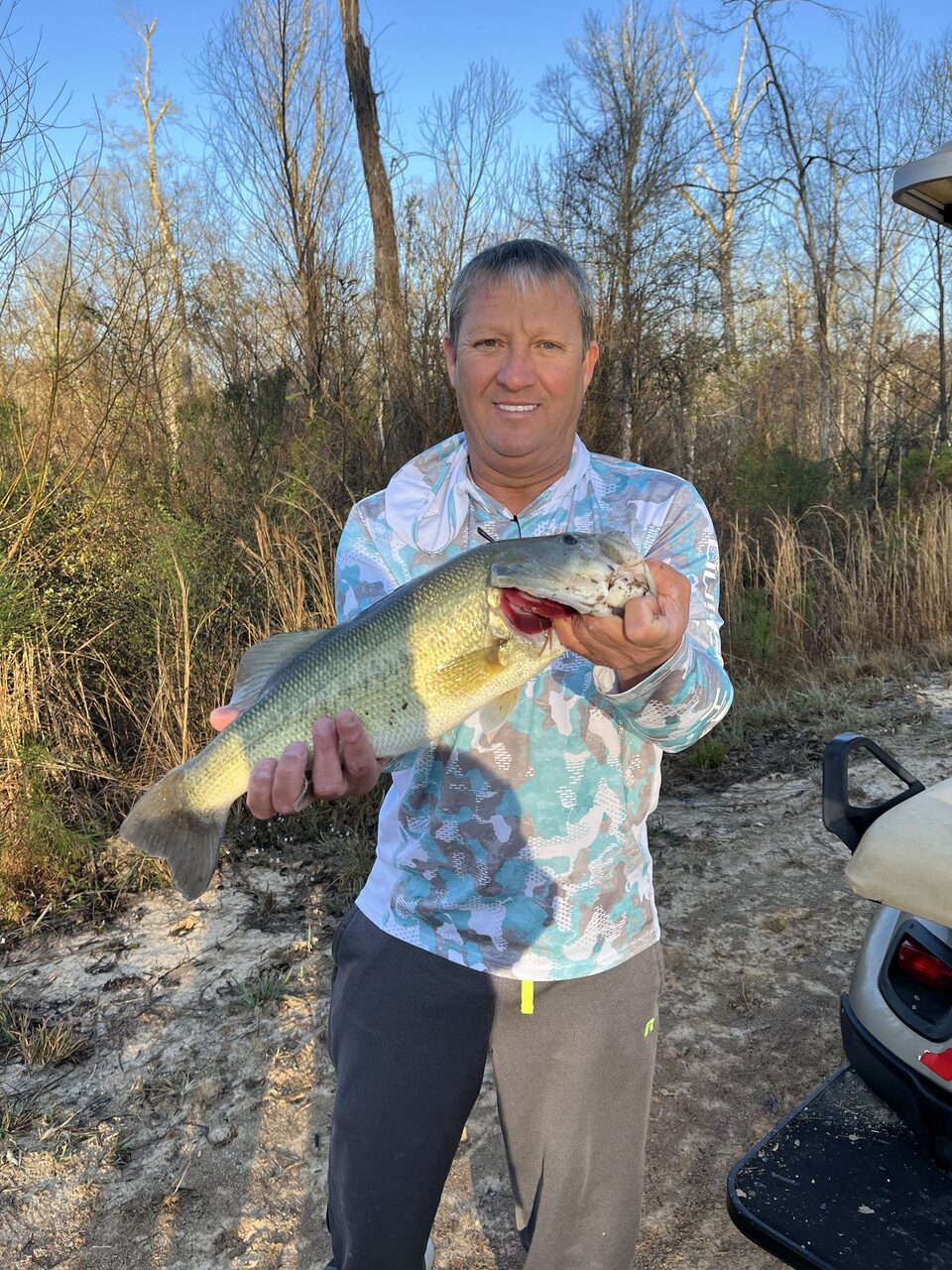 Smiling man in a long-sleeve fishing shirt holding up a largemouth bass under bare winter trees beside a golf cart on a sunny day