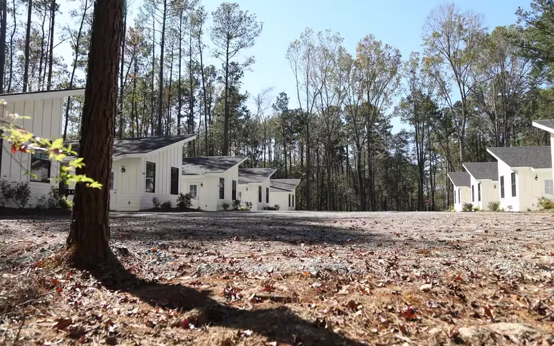 A row of modern, white cabins with dark shingled roofs sits in a clearing surrounded by tall pine trees at Fireside RV Resort in Ponchatoula, Louisiana.